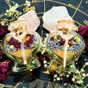 Könnte beinhalten: Zwei herzförmige Glasflaschen gefüllt mit Schichten aus getrockneten Blumen, Kräutern und Sand. Die Flaschen haben Korken und sind von getrockneten Rosen, Kristallen und Zweigen weißer Blumen umgeben. Der Hintergrund ist ein dunkler Stoff mit goldenen Himmelsmustern.