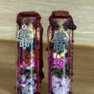 Könnte beinhalten: Zwei kleine Glasflaschen gefüllt mit getrockneten Blumen, Kristallen und anderen Pflanzenstoffen. Jede Flasche ist mit einem Korken und dunkelrotem Wachs versiegelt. Ein silberner Hamsa-Hand-Anhänger hängt von der Oberseite jeder Flasche.