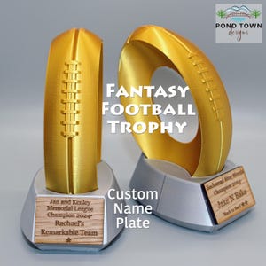 Puede incluir: Dos trofeos de fútbol de fantasía dorados con forma de balón de fútbol. Cada trofeo tiene una base plateada y una placa de nombre de madera. Se muestra el texto "FANTASY FOOTBALL TROPHY". Una placa dice "Jan and Kenley Memorial League Champion 2024- Rachael's Remarkable Team".