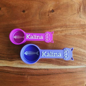 May include: Two purple and blue cat-shaped measuring spoons with a fishbone design and the name "Kalina" printed on them.