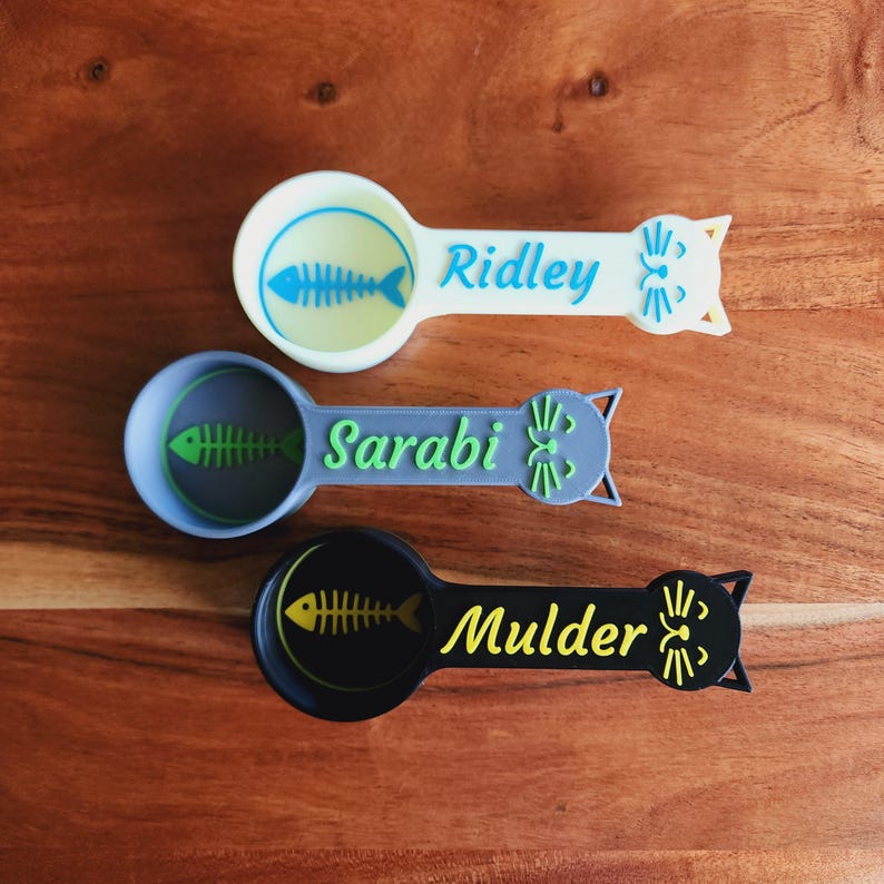 May include: Three white, gray, and black cat-shaped measuring spoons with a fishbone design and the names "Ridley", "Sarabi", and "Mulder" printed on them.