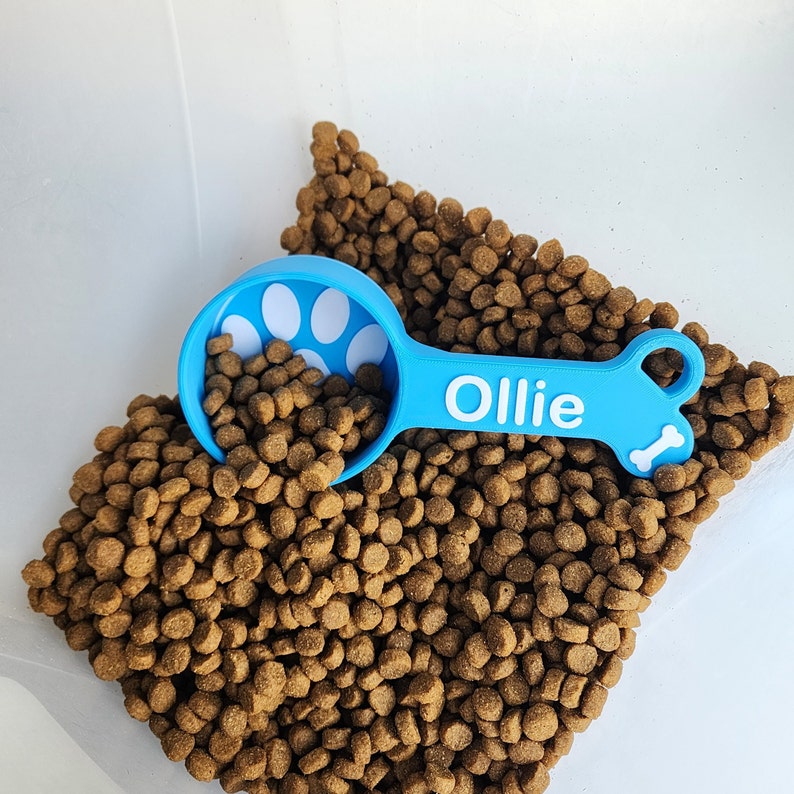 A light blue 1/2 cup dog food scoop with white accent for Ollie sitting in a container of dog food, the scoop is half filled with dog food