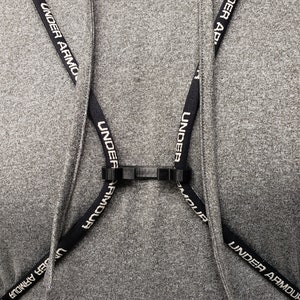 May include: A black strap with the text "Under Armour" printed on it is attached to a black plastic clip. The strap is crossed over itself and attached to another black plastic clip. The strap is worn over a gray hooded sweatshirt.