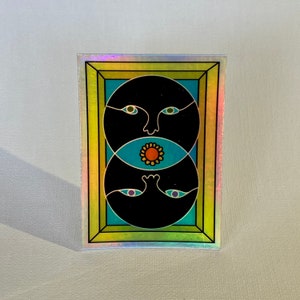 May include: A sticker with a psychedelic design featuring two black faces with blue eyes and a yellow flower in the center. The sticker is framed in a gold frame with a rainbow iridescent background.
