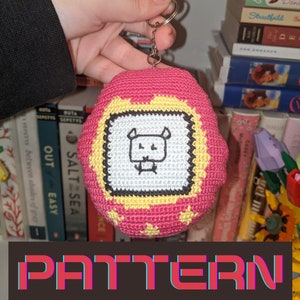 May include: A pink crocheted keychain featuring a retro video game design. The keychain has a white and yellow border with a black and white graphic. The word "CROCHET" and "PATTERN" are displayed in the image.