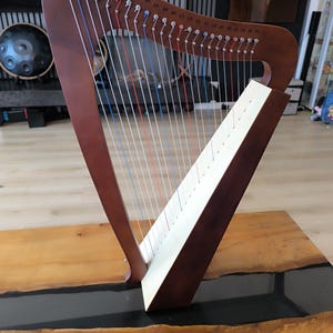 Op de afbeelding: Een bruine houten harp met snaren in verschillende kleuren, waaronder rood, blauw en wit. De harp staat op een houten tafel met een donker, glanzend midden. De snaren van de harp zijn gelabeld met muzieknoten.