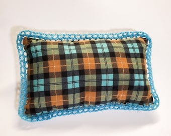 May include: A rectangular pillow with a blue, green, black, and orange plaid pattern. The pillow is trimmed with a blue crocheted border.