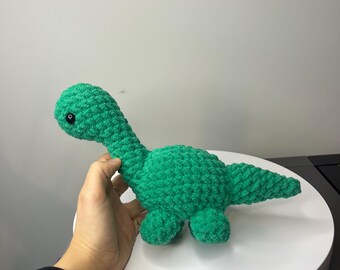 May include: A green crocheted dinosaur toy with black eyes. The dinosaur is sitting on a white surface.