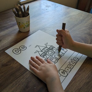 May include: A child's hand coloring a black and white illustration of a castle with a brown pencil. The illustration is on a white sheet of paper with the letter "C" and the word "Castle" written on it.