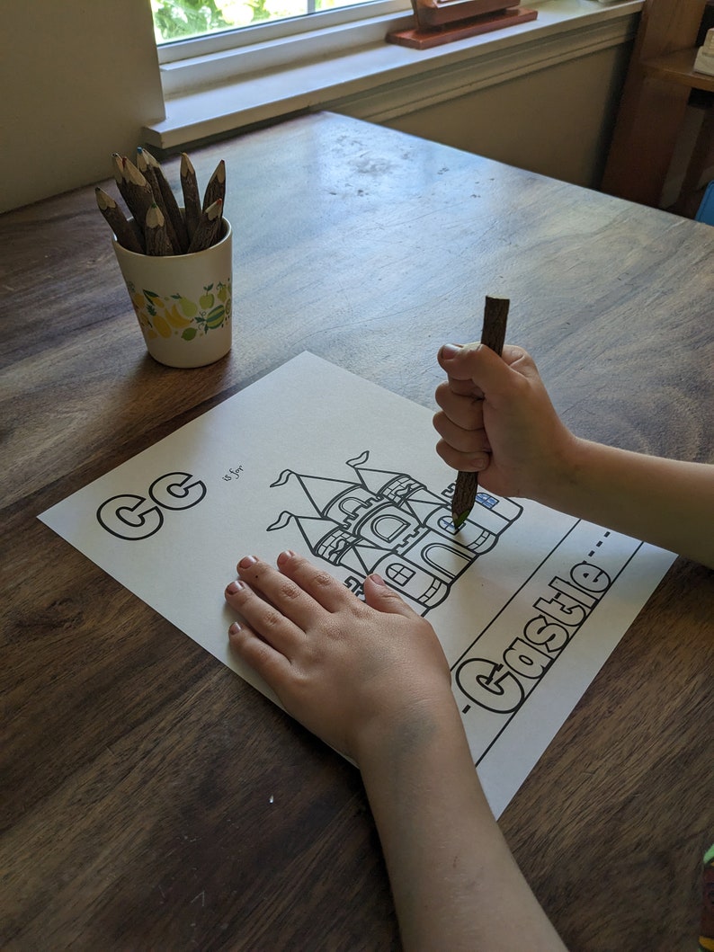 May include: A child's hand coloring a black and white illustration of a castle with a brown pencil. The illustration is on a white sheet of paper with the letter "C" and the word "Castle" written on it.