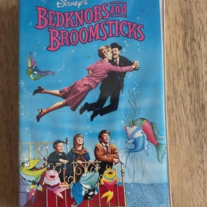May include: A VHS tape of Disney's "Bedknobs and Broomsticks" with a blue background and colorful illustrations. The cover features characters dancing and swimming with fish. The title is in pink and purple lettering.
