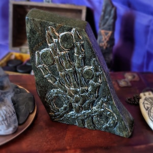 May include: A dark green stone sculpture of a hand with intricate details. The sculpture is carved with a raised design of a hand with fingers and a wrist.