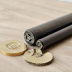May include: A collection of metal stamps and handles. Two cylindrical black handles are positioned behind two circular stamps and a third stamp. The stamps feature intricate Celtic and geometric designs, with a gold and silver finish.