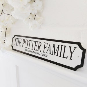 May include: A white sign with black trim and text that reads "THE POTTER FAMILY" with the names Mike, Janice, Luke, Sam & Charlie below. The sign is mounted on a white wall, with white flowers in the background.