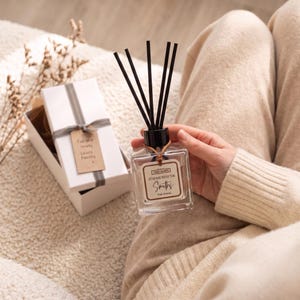 May include: A clear glass reed diffuser with black reeds and a label reading AT HOME WITH THE SMITHS Park Avenue. Beside it sits a white gift box with a grey ribbon and a tag inscribed with For the lovely Lewis Family x.