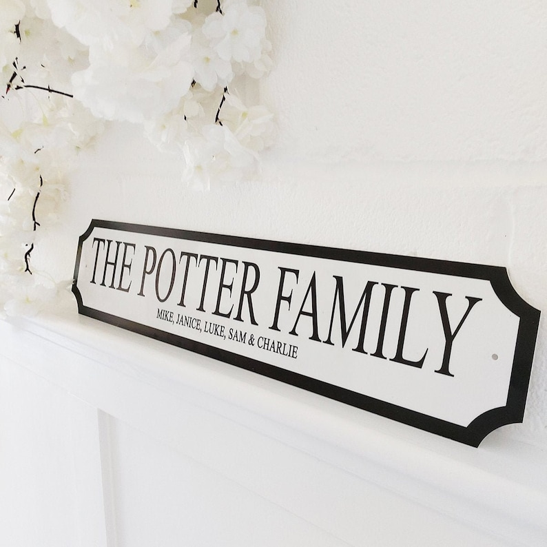 May include: A white sign with black trim and text that reads "THE POTTER FAMILY" with the names Mike, Janice, Luke, Sam & Charlie below. The sign is mounted on a white wall, with white flowers in the background.