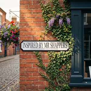 May include: A brick wall with a sign that reads "INSPIRED BY MR SHAPPERS" and a hanging basket of colorful flowers. The sign also says "A HAND CRAFTED DEVELOPMENT MADE IN LEICESTERSHIRE". Green vines and purple flowers are growing on the wall.