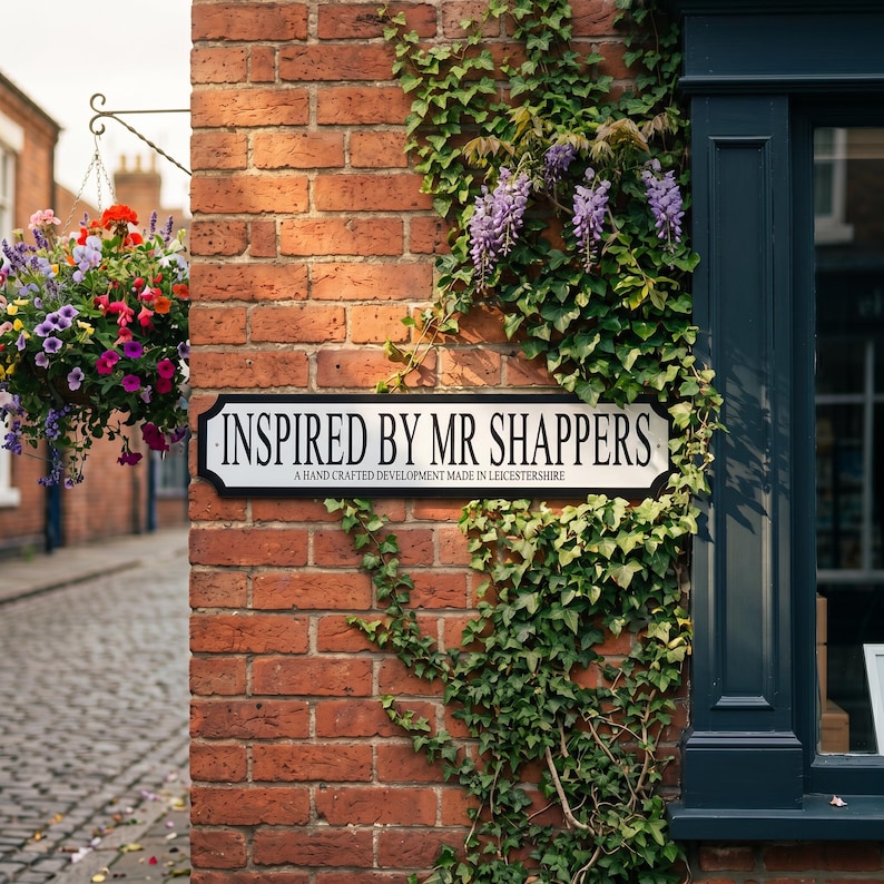 May include: A brick wall with a sign that reads "INSPIRED BY MR SHAPPERS" and a hanging basket of colorful flowers. The sign also says "A HAND CRAFTED DEVELOPMENT MADE IN LEICESTERSHIRE". Green vines and purple flowers are growing on the wall.