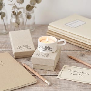 May include: A white ceramic tea cup shaped candle holder with a lit tea light inside. The cup features a black line drawing of a teapot and branches with the text "Thank you for being amazing." It sits on a matching tan cardboard gift box.
