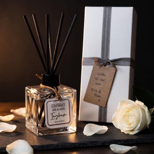 May include: A square glass reed diffuser with black sticks and a label reading MR & MRS Ingham Adam & Olivia. Next to it is a white gift box with a grey ribbon and a brown tag reading With all our love Ella & Tom x, plus a white rose.