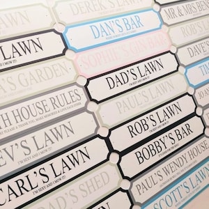 May include: A collection of decorative signs with various names and phrases. The signs are rectangular with a white background and black borders. Some signs have colored accents, such as blue or pink. Text includes "Dan's Bar" and "Dad's Lawn."