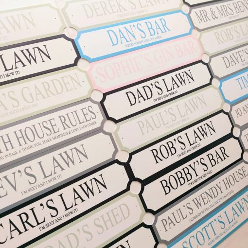 May include: A collection of decorative signs with various names and phrases. The signs are rectangular with a white background and black borders. Some signs have colored accents, such as blue or pink. Text includes "Dan's Bar" and "Dad's Lawn."