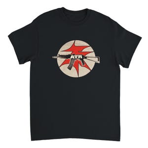 Puede incluir: Una camiseta negra de manga corta con un gráfico circular en el pecho. El diseño incluye un círculo beige con una forma roja de estrella detrás de la silueta de un rifle negro. Las letras ATR están impresas en blanco sobre el rifle.