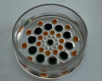 May include: Clear glass coaster with a circular design of green, orange, and silver dots. The coaster has a raised edge and a flat base. The dots are arranged in a spiral pattern, creating a decorative and functional item.