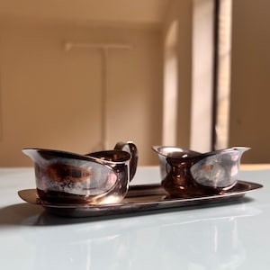 May include: A silver-toned creamer and sugar bowl set. The creamer has a handle and a curved spout. The sugar bowl is a smaller version of the creamer. Both sit on a matching oval tray. The set is reflective and sits on a light blue surface.
