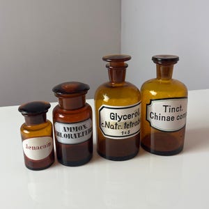 Peut inclure: Quatre flacons de pharmacie anciens en verre ambré avec des bouchons brun foncé. Chaque flacon porte une étiquette blanche avec du texte noir. Les étiquettes indiquent "Jenacain", "Ammon. Chlorat. Fer", "Glycerol c. Natr. tetrabor" et "Tinct. Chinae com".