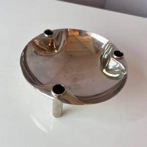 Könnte beinhalten: Ein silberfarbene, kreisförmige Dekoschale mit drei schwarz ausgekleideten Löchern und einem einzelnen zylindrischen Bein. Die Schale hat eine reflektierende Oberfläche und ein geschwungenes Design.