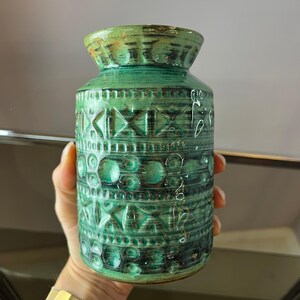 May include: A green ceramic vase with a textured surface and geometric patterns. The vase has a cylindrical shape with a slightly flared top. The color is a mix of green and brown, with a glossy finish. The vase is approximately 8 inches tall.