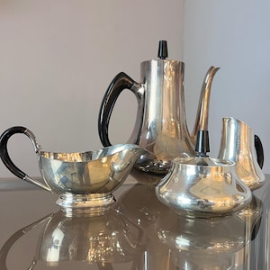 May include: A silver tea set with black handles and accents. The set includes a teapot, creamer, sugar bowl, and a gravy boat. The reflective surface adds to the elegance of the set.