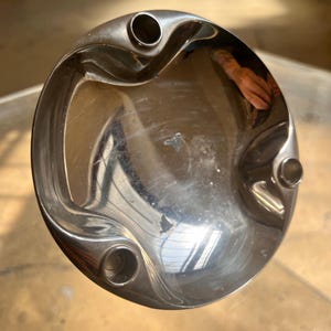 Può includere: Un posacenere circolare in metallo lucido. Il posacenere ha una forma a ciotola poco profonda con tre rientranze circolari per tenere le sigarette. La superficie riflette l'ambiente circostante.