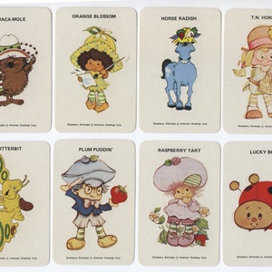Op de afbeelding: Een set van acht Strawberry Shortcake-personagekaarten. Elke kaart heeft een ander personage met een unieke naam en een kleurrijke illustratie. De personages zijn Guaca-Mole, Orange Blossom, Horse Radish, T.N. Honey, Flitterbit, Plum Puddin', Raspberry Tart en Lucky Bug.