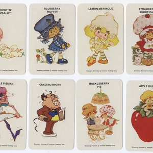 Op de afbeelding: Een set van acht Strawberry Shortcake-personagekaarten. Elke kaart toont een ander personage met zijn naam en een korte beschrijving. De personages zijn: Apricot 'n' Hopsalot, Blueberry Muffin, Lemon Meringue, Strawberry Shortcake, Purple Pieman, Coco Nutwork, Huckleberry Pie en Apple Dumplin'.