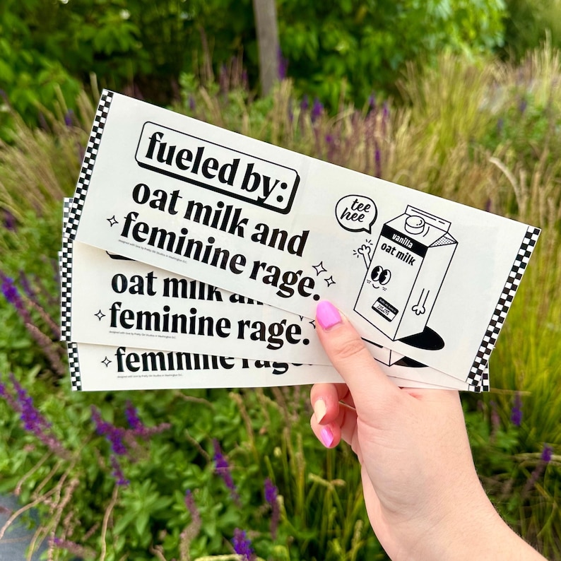 Puede incluir: Una pegatina rectangular blanca con un borde de cuadros negros y el texto "fueled by: oat milk and feminine rage." junto a una ilustraci&oacute;n de un cart&oacute;n de leche de avena de vainilla con un bocadillo que dice "tee hee" en tinta negra.