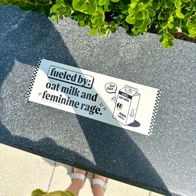 Puede incluir: Una pegatina rectangular blanca con un borde de cuadros negros. El texto dice "fueled by: oat milk and feminine rage". Incluye un dibujo animado de un cart&oacute;n de leche de avena con cara, brazos y un bocadillo que dice "tee hee".