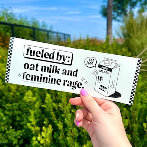 Puede incluir: Una pegatina rectangular blanca con un borde de tablero de ajedrez negro en los lados izquierdo y derecho. El texto dice fueled by: oat milk and feminine rage. Un cart&oacute;n de leche de avena con cara y brazos est&aacute; junto a una burbuja que dice tee hee.