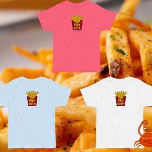May include: Three t-shirts featuring a graphic of french fries in a red container with the text "Fry Day!" on each shirt. The shirts are pink, light blue, and white.