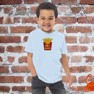 May include: A light blue t-shirt with a red and yellow graphic of french fries in a red container with the text "Fry Day!" printed on it.
