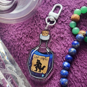 Peut inclure: Un charme en forme de bouteille en verre transparent avec un bouchon en liège et un liquide bleu à l'intérieur. La bouteille porte une étiquette sur laquelle est inscrit "Loup-Garou" avec une illustration de loup-garou noir. Le charme est attaché à un porte-clés en métal blanc.