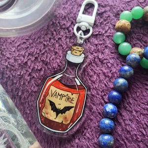 Può includere: Un portachiavi in acrilico trasparente con un design a forma di bottiglia di pozione. La bottiglia contiene liquido rosso e un'etichetta con la scritta "Vampire" e un'illustrazione di pipistrello. Con chiusura bianca.