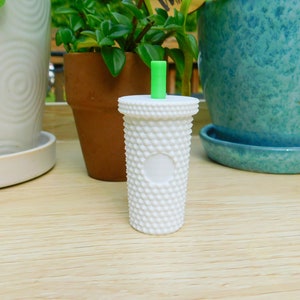 May include: White ceramic cup with a textured surface and a green straw. The cup has a white circle in the center.