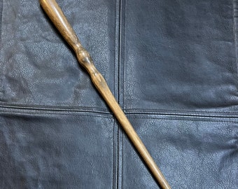 May include: A wooden wand with a dark brown finish and a slightly curved handle. The wand is lying on a dark brown leather surface.