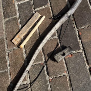 May include: A set of wooden tools including a long, weathered gray wooden rod with a black cord, a small rectangular gray wooden block with a central hole, a light-colored wooden dowel, and a flat, notched rectangular wooden piece.