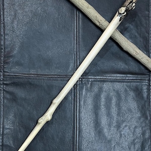 May include: A smooth, light-colored wooden wand featuring a tapered shape and a natural knot near the bottom. The handle end is darkened with a charred, textured finish. A second, rougher, unrefined wooden stick lies diagonally across the wand.