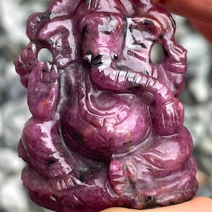 May include: A carved, deep magenta gemstone figurine of Ganesha, the Hindu deity. The sculpture features intricate details, including the elephant head, multiple arms, and seated posture. The gemstone has a polished, reflective surface.