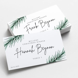 Peut inclure: Deux cartes de place blanches avec des illustrations de branches de pin vertes. La carte du haut indique "MILLERS CHRISTMAS DINNER Frank Beynon TABLE 2". La carte du bas indique "MILLERS CHRISTMAS DINNER Hannah Beynon TABLE 1 24.12.2021".