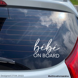 May include: White vinyl car sticker with the text "bébé ON BOARD" in a cursive font. The sticker is on the rear window of a car.